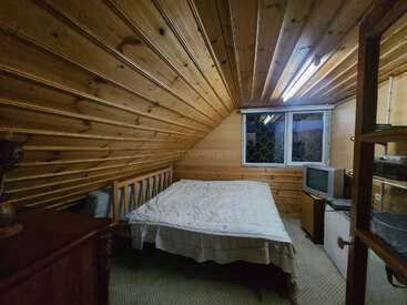 Esta acogedora habitación en el ático cuenta con paredes y techo inclinados de madera, una cama doble, una ventana pequeña, luz fluorescente, estanterías y un televisor antiguo.