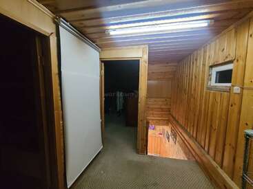 Esta imagen muestra un pasillo de madera con suelo enmoquetado, iluminación fluorescente, una puerta que da a una habitación más oscura, unas escaleras, una ventana pequeña y un panel blanco.