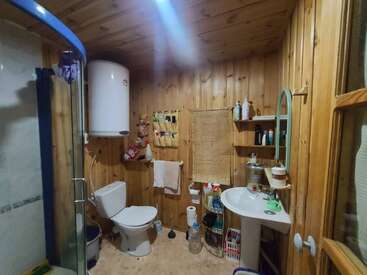 Este acogedor cuarto de baño cuenta con paredes y techo de madera, un inodoro, un lavabo con espejo, una cabina de ducha, un calentador de agua, estantes con artículos de aseo y diversos objetos personales.