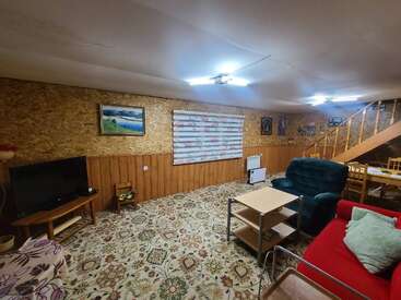 Esta acogedora habitación cuenta con paredes revestidas de madera, moqueta estampada, un televisor, sofás verdes y rojos, mesitas, una iluminación brillante y unas escaleras que suben. Está decorada con cuadros.