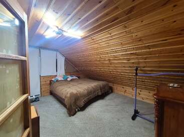 Esta acogedora habitación en el ático cuenta con paredes y techo de madera, una cama con ropa de cama marrón, suelo enmoquetado, un perchero, una cómoda, lámparas de techo luminosas y persianas.