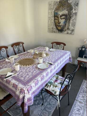 Hay una mesa puesta para cinco personas con platos y tazas blancas, cubierta con un mantel morado estampado. En la pared cuelga un cuadro de Buda.