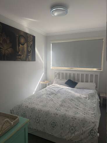 Una acogedora habitación con una cama blanca, un edredón estampado, mesitas de noche pequeñas, persianas cerradas, una tranquila obra de arte de Buda, un baúl verde menta y una suave luz natural que se filtra por la ventana.