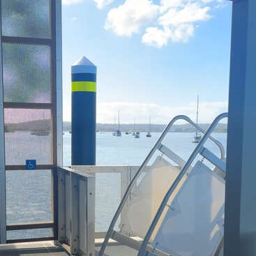 Das Bild zeigt eine sonnige Uferlandschaft mit Booten, die in der Ferne vor Anker liegen, einem hohen blau-gelben Stegpfosten, Metallgeländern und einem Schild, das auf die Barrierefreiheit für Rollstuhlfahrer hinweist.