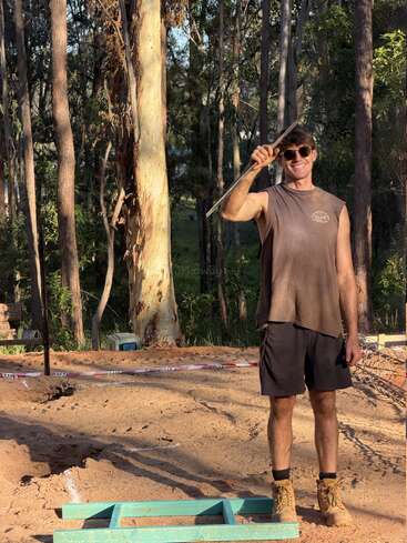 Un hombre con gafas de sol, camiseta sin mangas y botas de trabajo está de pie sobre un terreno arenoso, sosteniendo una herramienta metálica y sonriendo, rodeado de árboles altos bajo la luz del sol.