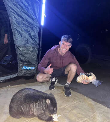 Un hombre está sentado en cuclillas junto a una tienda de campaña por la noche, sonriendo y levantando el pulgar, mientras sostiene un trozo de pan. Un wombat come pan sobre una estera cerca de allí.