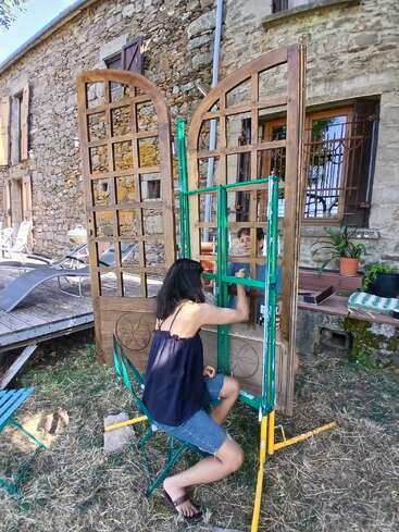 Eine Frau sitzt auf einem Stuhl und streicht ein großes Holztor vor einem rustikalen Steinhaus, umgeben von Grünpflanzen und Gartenmöbeln an einem sonnigen Tag.