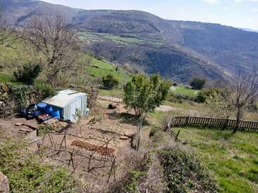 Ein Garten am Hang mit einem Schuppen, blauen Fässern, einem Holzzaun, Beeten und vereinzelten Bäumen, von dem aus man einen malerischen Blick auf grüne Berge und das darunter liegende Tal hat.