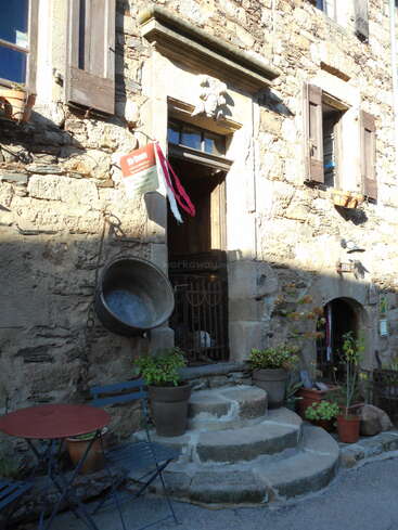 La imagen representa la fachada de un edificio de piedra con un portal, ventanas y macetas, con un cartel y una gran maceta metálica colgados de la pared.
