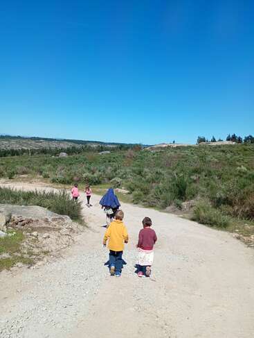 Five children walk along a dirt path in a wide, open landscape with green bushes under a bright blue sky on a sunny day outdoors.