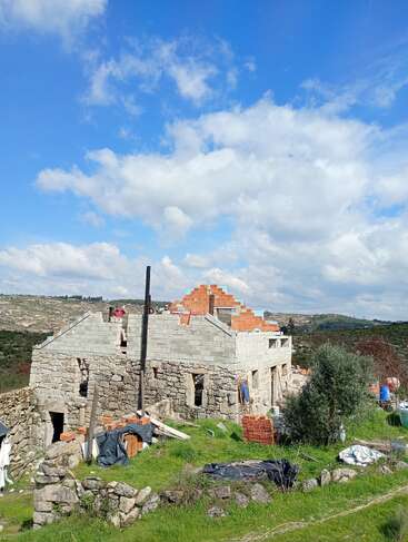 A stone house under construction stands on a grassy hill, surrounded by building materials. Blue sky with fluffy clouds forms a striking backdrop over rural landscape.