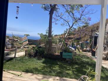 A backyard garden with green grass, trees, and scattered outdoor furniture. There are gardening tools, a shed, and a view of the sea in the background.