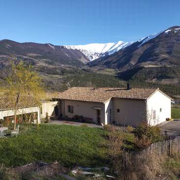Une charmante maison de campagne au toit de tuiles se dresse au milieu d'une pelouse verdoyante, au pied de majestueuses montagnes enneigées et sous un ciel bleu limpide, offrant un paysage paisible et pittoresque.