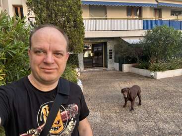 Um cara tira uma selfie do lado de fora de um prédio com um cachorro atrás dele numa área pavimentada. No fundo, dá pra ver vegetação e varandas.