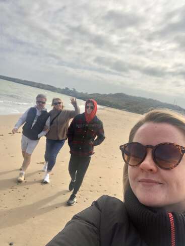 A imagem mostra uma selfie de uma mulher em uma praia com três pessoas atrás dela, usando roupas casuais e óculos escuros, com um céu nublado e colinas ao fundo.