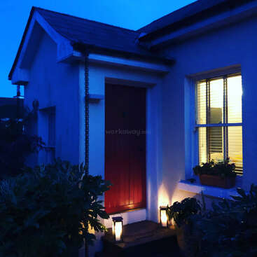 A imagem mostra uma casa com uma porta vermelha, uma janela com plantas e uma pequena varanda iluminada por lanternas ao anoitecer.