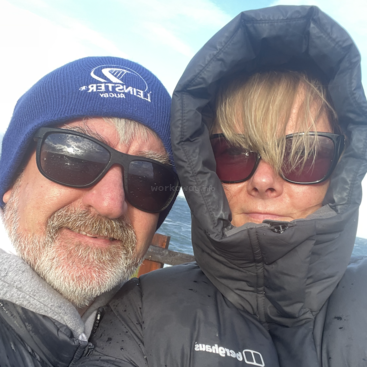 A imagem mostra um homem e uma mulher juntos ao ar livre, usando roupas de inverno e óculos escuros, com um corpo de água visível ao fundo.