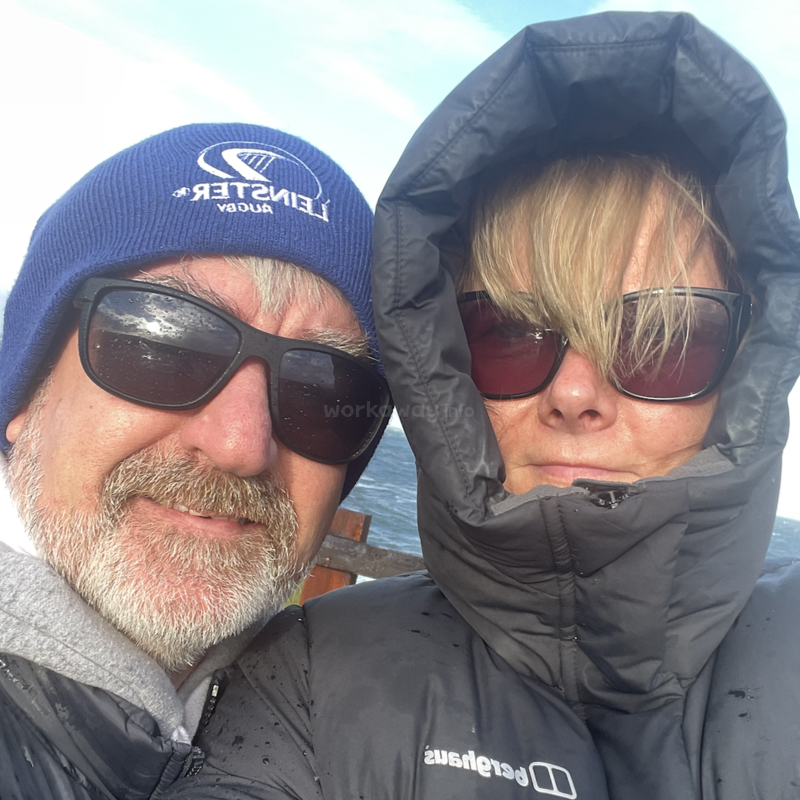 A imagem mostra um homem e uma mulher juntos ao ar livre, usando roupas de inverno e óculos escuros, com um corpo de água visível ao fundo.