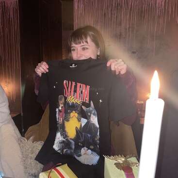 Une femme brandit joyeusement un t-shirt "Salem Moon" représentant diverses images de chats. Il y a une bougie allumée, des cadeaux de Noël et des traces de peinture rose sur le mur.