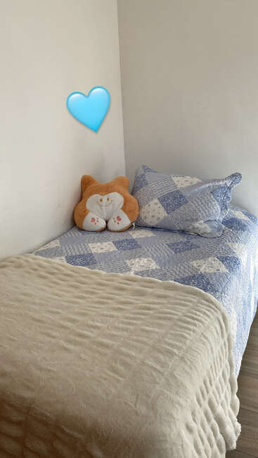 A cozy, neatly made single bed with a blue patchwork quilt, fluffy beige blanket, cat-shaped plush pillow, and a blue heart emoji on the wall.