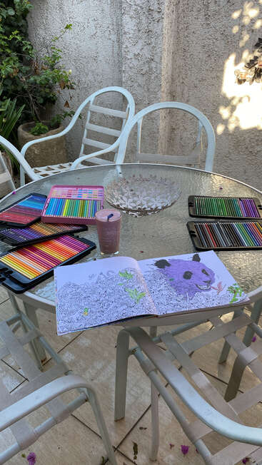 A glass patio table holds colored pencils, a coloring book featuring a purple panda, a smoothie, and an empty bowl, surrounded by white chairs and garden plants.