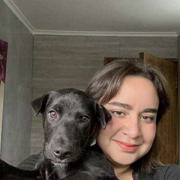 A woman with brown hair smiles warmly while holding a black dog. Both appear happy, posing together indoors near a wooden door in a cozy room.