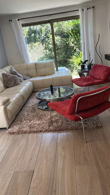 Uma sala de estar moderna e aconchegante com um sofá seccional creme, duas cadeiras vermelhas, mesa de centro de vidro, tapete felpudo, piso de madeira e vista para um jardim exuberante.