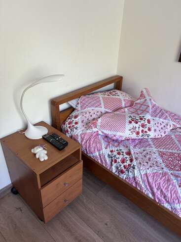Um quarto pequeno e aconchegante com uma cama de madeira, colcha de retalhos floral rosa, mesa de cabeceira, abajur branco, dois controles remotos e um ursinho de pelúcia pequeno.