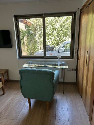 Um quarto aconchegante com uma poltrona verde de frente para uma mesa de vidro, janela grande, piso de madeira e armário, e vista para um jardim e um carro estacionado.