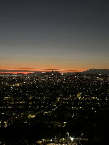 Uma bela paisagem urbana ao entardecer, com as luzes cintilantes da cidade, arranha-céus ao longe, montanhas recortadas contra o céu e um horizonte alaranjado e vibrante sob um céu noturno claro que vai escurecendo.