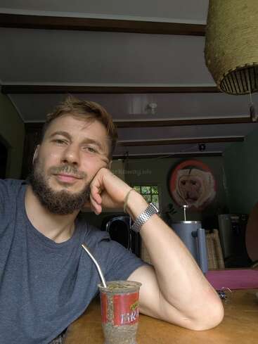 Ein bärtiger Mann sitzt in einem Haus, stützt sein Gesicht auf seine Hand und trinkt Yerba Mate. Hinter ihm sind ein Wandgemälde mit einem Affen und verschiedene Gegenstände zu sehen.
