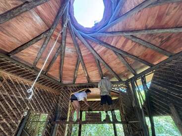 Two people work inside a partially built wooden hut with a circular roof opening above. The lattice walls and natural wood beams create a rustic structure.