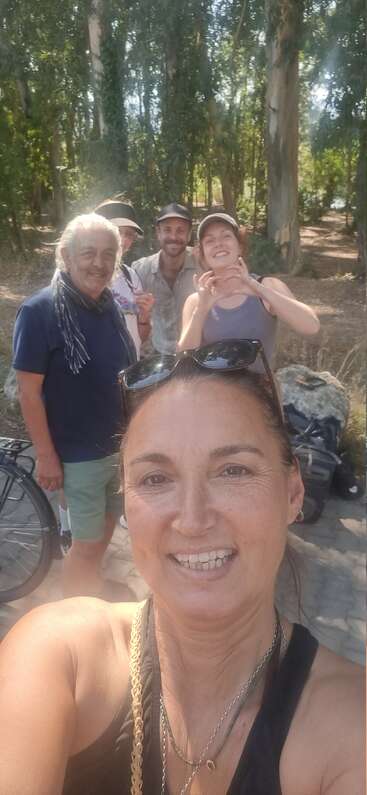 Un groupe de cinq personnes souriantes prend un selfie en plein air, entouré de grands arbres, de la lumière du soleil et de la nature, profitant d'une agréable journée ensemble, avec des vibrations détendues.