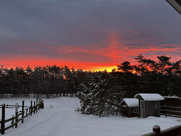 Un paysage enneigé met en scène une clôture en bois, un chalet et des arbres recouverts de neige. Derrière la forêt, un coucher de soleil rouge et orange éclatant illumine le ciel nuageux spectaculaire.