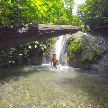 A woman in a bikini stands in a shallow pool of water, facing a small waterfall, surrounded by lush greenery and large rocks.