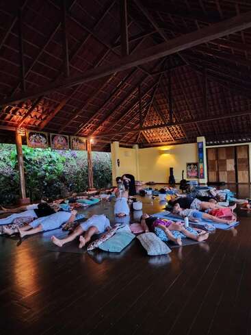 A group of people lie relaxed on yoga mats and cushions inside an open wooden pavilion, engaging in a calming meditation or sound healing session, surrounded by nature.