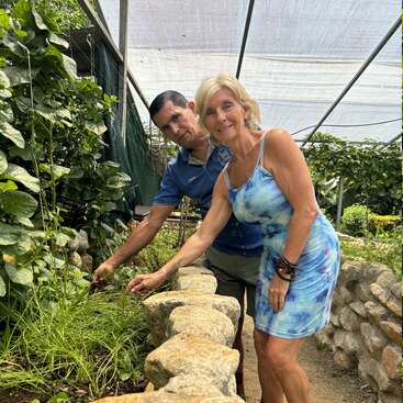 A man and woman are gardening together in a greenhouse, smiling and tending to plants. Stone walls and lush greenery surround them under a netted canopy.