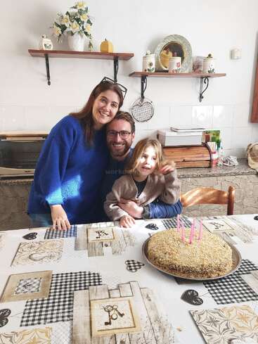 Eine dreiköpfige Familie lächelt um einen Tisch mit einem Geburtstagskuchen, der mit vier rosa Kerzen gekrönt ist, und feiert gemeinsam in einer gemütlichen, warm dekorierten Küche.