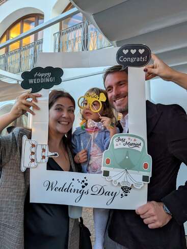 Eine glückliche Familie posiert freudig lächelnd mit einem Fotorahmen zum Thema Hochzeit. Das Kind trägt eine verspielte "Braut"-Brille. Auf den dekorativen Schildern steht "Happy Wedding" und "Congrats!".