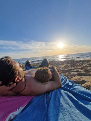 Ein Mann und ein Kind liegen auf Strandtüchern, entspannen sich gemeinsam und blicken bei Sonnenuntergang auf das Meer.