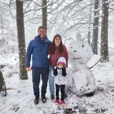 Eine dreiköpfige Familie, warm angezogen, steht lächelnd in einem verschneiten Wald neben einem großen, kreativen Schneemann. Der Schnee bedeckt den Boden, die Bäume und die Äste.