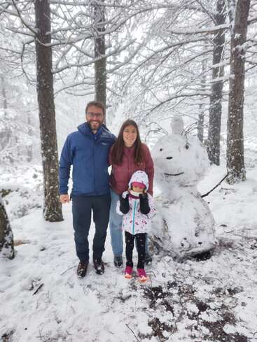 A family of three, warmly dressed, stands smiling in a snowy forest beside a large, creative snowman. Snow covers the ground, trees, and branches.