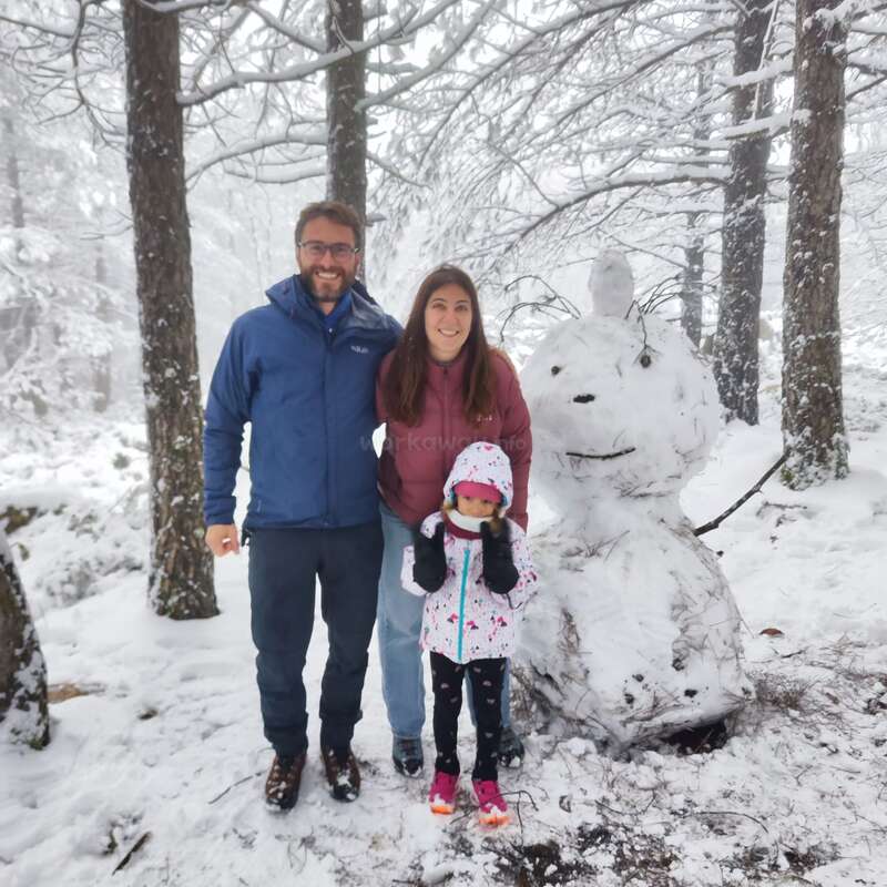 Eine dreiköpfige Familie, warm angezogen, steht lächelnd in einem verschneiten Wald neben einem großen, kreativen Schneemann. Der Schnee bedeckt den Boden, die Bäume und die Äste.
