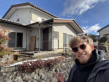 A smiling man wearing sunglasses poses with a peace sign in front of a traditional Japanese house on a sunny winter day, light snow visible on bushes.