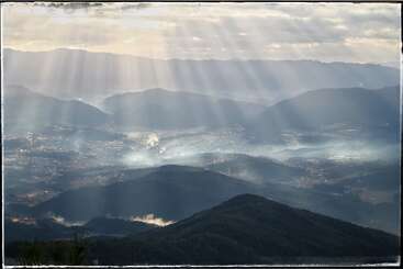 Golden sunlight streams through dramatic clouds, illuminating the misty mountain valley below. Soft rays highlight scattered smoke, creating a peaceful, ethereal scene of natural beauty and tranquility.