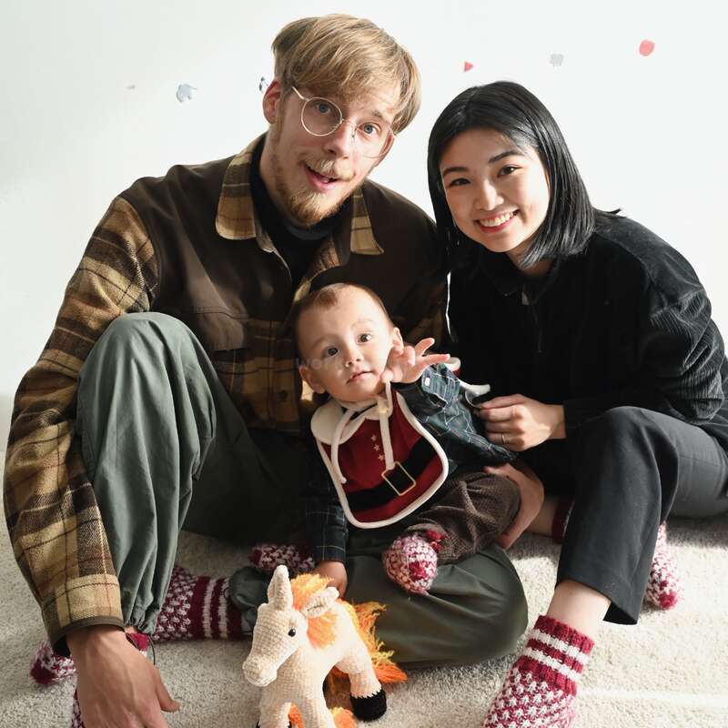 A happy family of three sits on the floor: a smiling man, woman, and baby with a unicorn toy. Cozy atmosphere, colorful woolen socks.