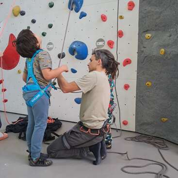 Un niño, equipado con un arnés de escalada, recibe ayuda de un adulto en un rocódromo cubierto. La pared que tienen detrás está cubierta de presas de colores.