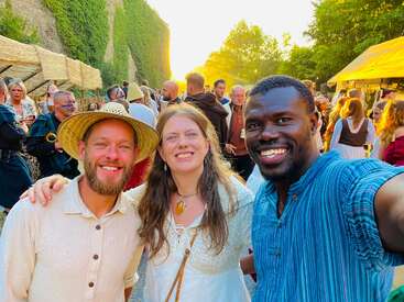 Three friends smile at a lively outdoor festival, dressed in medieval attire. The crowd behind them mingles under warm sunlight, creating a joyful, festive atmosphere.