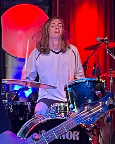 A young drummer passionately performs on stage, eyes closed, lost in music. Colorful lights illuminate the scene, highlighting his energy and the dynamic atmosphere around him.