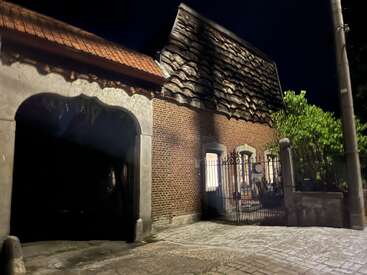 The image shows a brick house with a unique, wavy roof, lit at night. There’s an arched gate, iron fence, trees, and a streetlamp nearby.